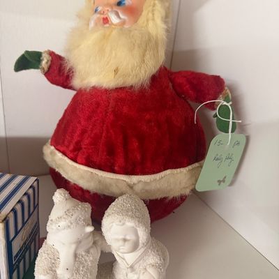 Vintage roly-poly plush Santa Claus doll featuring a painted rubber face and fuzzy beard, displayed alongside a white snow-covered figurine.