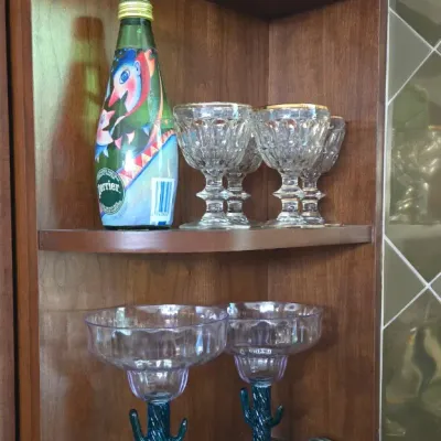 Sale Photo Thumbnail #9: Collection of glassware displayed on a wooden wall shelf, including novelty cactus-stem margarita glasses, gold-rimmed crystal goblets, and an art-labeled Perrier bottle.
