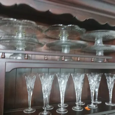 Sale Photo Thumbnail #22: Collection of clear pressed glass pedeled cake stands and cut glass champagne flutes displayed on a dark wood china hutch shelf.