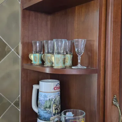 Sale Photo Thumbnail #11: Collection of glassware displayed on open wooden shelves, including vintage Coors and Olympics mugs, shot glasses with decorative holders, and textured tall glasses.