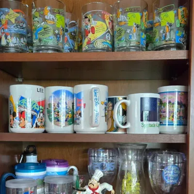 Sale Photo Thumbnail #15: Collection of vintage Disney souvenir drinkware displayed on cabinet shelves, including printed drinking glasses, mugs, acrylic tumblers, and an Epcot chef figurine.