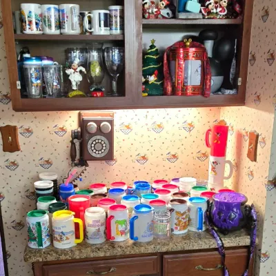Sale Photo Thumbnail #16: Large collection of Disney souvenirs, including refillable resort mugs, glassware, figurines, and holiday decor displayed on a desk and in a wooden wall cabinet.