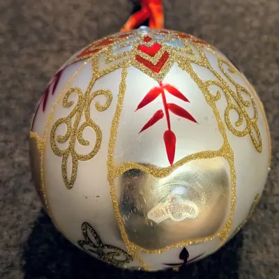 Sale Photo Thumbnail #80: Waterford glass Christmas ornament featuring intricate gold glitter scrolls, red accents, and the original Waterford sticker on a silver-white background.