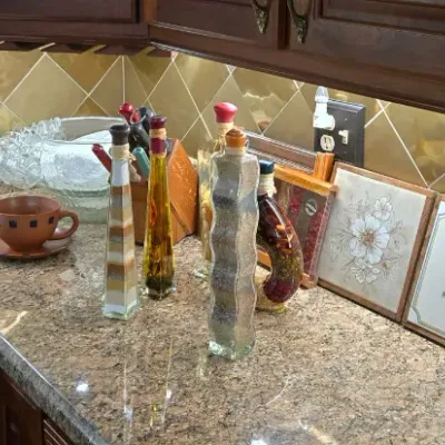 Sale Photo Thumbnail #7: Collection of kitchen decor and glassware arranged on a granite countertop, including decorative olive oil bottles, sand art bottles, and framed floral trivet tiles.