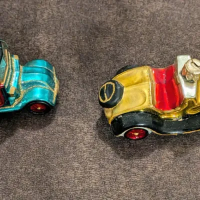 Sale Photo Thumbnail #79: Pair of vintage blown-glass Christmas ornaments shaped like antique automobiles, featuring metallic finishes and glitter accents.