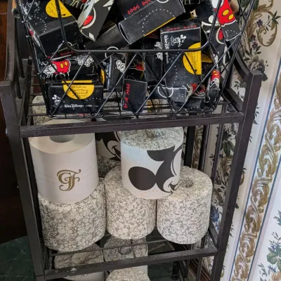 Sale Photo Thumbnail #88: Black metal shelving unit filled with vintage Walt Disney World Resort souvenir toilet paper rolls and boxed bath soaps.