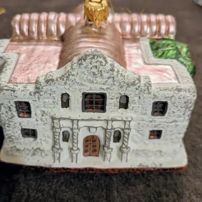 Sale Photo Thumbnail #73: Blown glass Christmas tree ornament shaped like the Alamo, featuring a textured white facade with metallic copper roof accents.