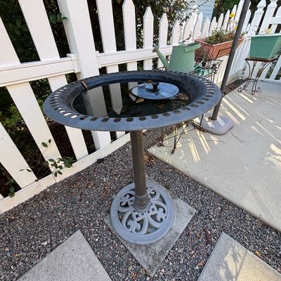 Cast metal pedestal birdbath featuring an ornate scrollwork base, slotted rim, and a small solar-powered fountain insert.