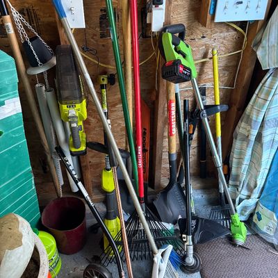 Collection of lawn and garden tools, including Ryobi cordless string trimmers, edger, vacuum, and various hand rakes, shovels, and brooms.