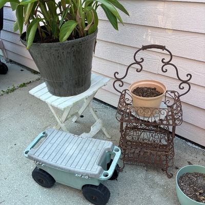 Collection of outdoor garden items including a rolling garden scooter, potted plants, a white wooden plant stand, and a vintage wrought...
