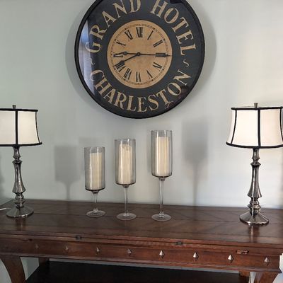Large Grand Hotel Charleston wall clock displayed above a wooden console table with a pair of metal table lamps and glass candle holders.