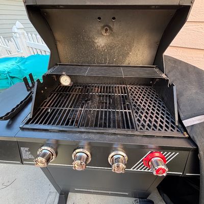 Black propane gas barbecue grill with open lid, featuring four front control knobs, an upper warming rack, and cast iron cooking grates.