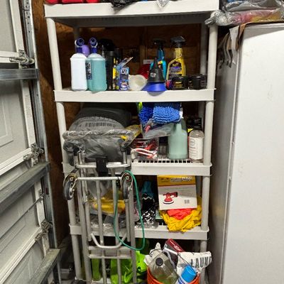 A five-tier white plastic shelving unit holding various garage supplies, car care products, and cleaning chemicals alongside a folding...
