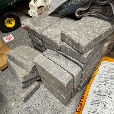 Stack of grey concrete patio pavers alongside gardening supplies.