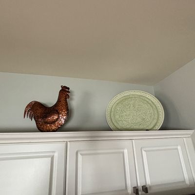 Hammered copper-tone rooster figurine alongside a pale green embossed ceramic platter, displayed above kitchen cabinetry.