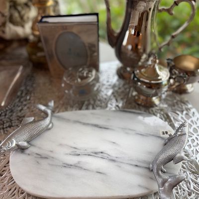White marble serving tray featuring detailed cast metal fish-shaped side handles, shown with silver-plated tea set items in the background.