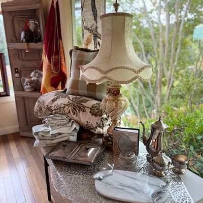 Tabletop display featuring a ornate cream table lamp with trim, silver-plated coffee and tea service, linens, pillows, and decorative...