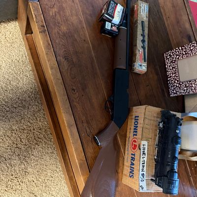 Vintage air rifle with faux wood stock resting on a wooden coffee table alongside a boxed scope, ammunition, and a vintage Lionel toy...