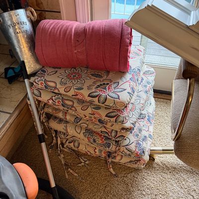 Stack of floral tie-on patio chair cushions topped with a rolled pink cushion, stored indoors next to miscellaneous household items.