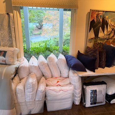 Collection of bed linens, decorative throw pillows, and pillow inserts displayed on folding tables next to a large abstract painting.