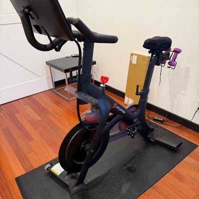 Peloton stationary exercise bike with attached touchscreen monitor, floor mat, and a pair of small weights mounted on the rear bracket.