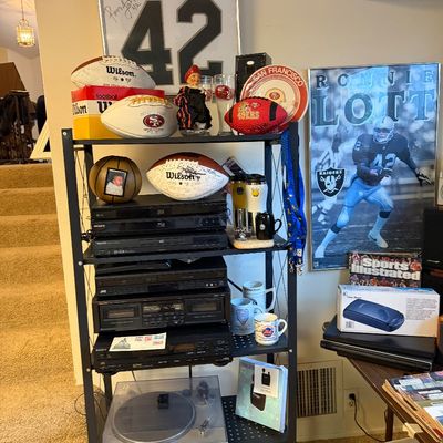 Metal shelving unit filled with vintage home audio equipment, home video players, and San Francisco 49ers sports memorabilia including...
