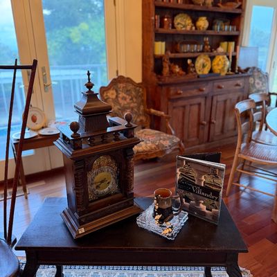 Antique wooden mantel clock displayed on a dark accent table alongside a coffee table book and a ceramic mug.