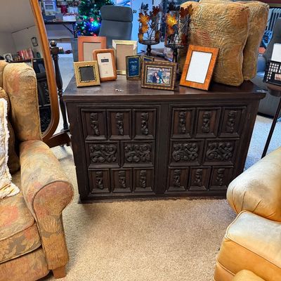 Vintage dark wood credenza featuring ornate carved panel doors, topped with an assortment of picture frames and decorative metal leaf...
