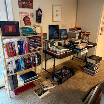 Large collection of hardcover and paperback books organized on a light wood bookshelf and a dark folding table, accompanied by framed...
