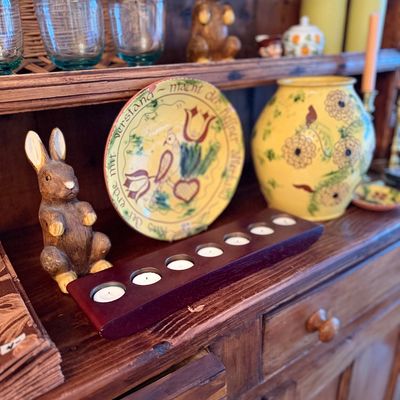 Rustic wooden hutch shelf featuring a hand-painted folk art decorative plate, a ceramic vase, rabbit figurines, and a dark wood votive...