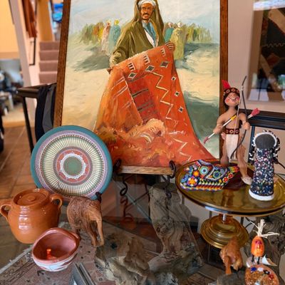 Framed oil painting depicting a rug merchant in traditional attire, displayed alongside folk art figurines, pottery, and decorative objects.