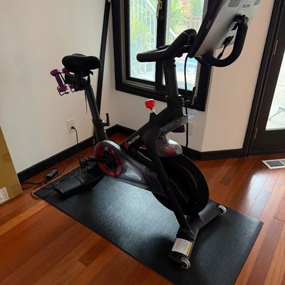 Peloton stationary exercise bike with attached touchscreen display, floor mat, and a pair of purple hand weights mounted on the rear.