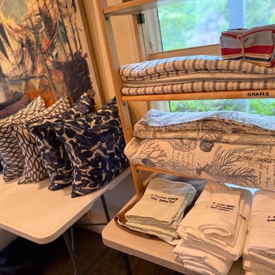 Collection of sorted bed linens and decorative throw pillows, including pillow shams, quilts, and coverlets arranged on shelves and tables.