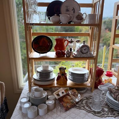 A light wood folding shelving unit displaying assorted tableware, including ceramic teapots, decorative plates, glassware, an orange...