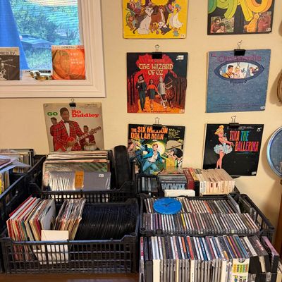 Collection of vintage vinyl records, CDs, and cassette tapes displayed in plastic crates and hung on the wall, including titles like...