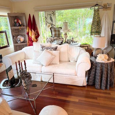 Sunlit living room featuring a white upholstered sofa with decorative pillows, a chrome glass-top coffee table, and various home decor...