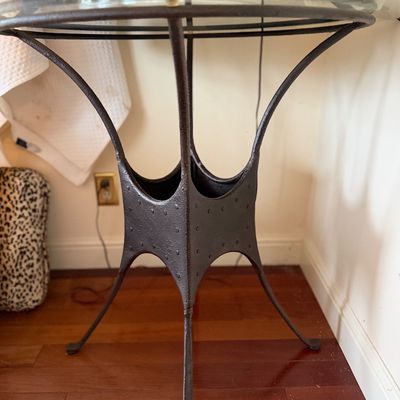 Round glass-top side table with a dark, textured cast iron or wrought metal base featuring studded accent panels.