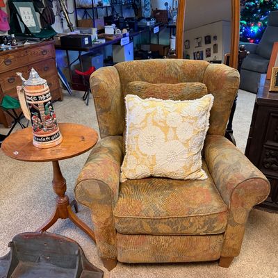 Upholstered floral armchair paired with a small round wooden pedestal side table holding a German beer stein, set alongside a cheval...