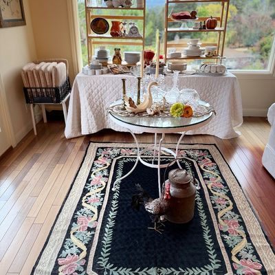 Black floral area rug featuring a glass-top accent table with crystal decor, flanked by a rustic milk can, a rooster figurine, and...