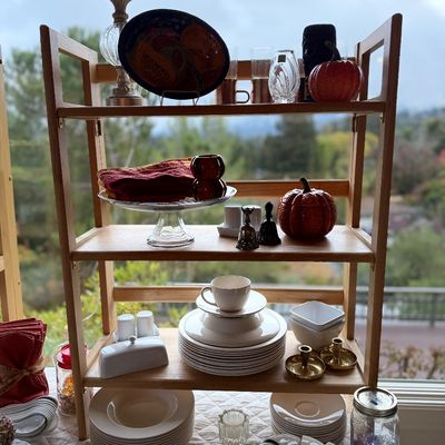 Three-tiered light wood shelving unit displaying fall decor, ceramic dishes, brass candlesticks, and glassware.