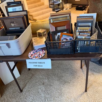 Brown laminate top folding table holding plastic crates and a tub filled with assorted picture frames and photo albums.