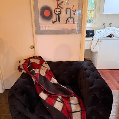 Black tufted velvet armchair draped with a red, black, and cream plaid wool blanket, positioned below a framed Joan Miró exhibition poster.