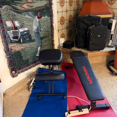 Weider weight bench alongside a black ergonomic kneeling chair, set on exercise mats near a woven golf tapestry hanging on the wall.