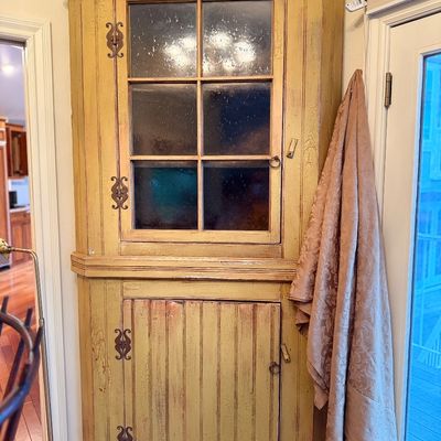 Rustic distressed yellow-painted corner hutch featuring a six-pane glass upper door, a beadboard lower cabinet door, and decorative iron...