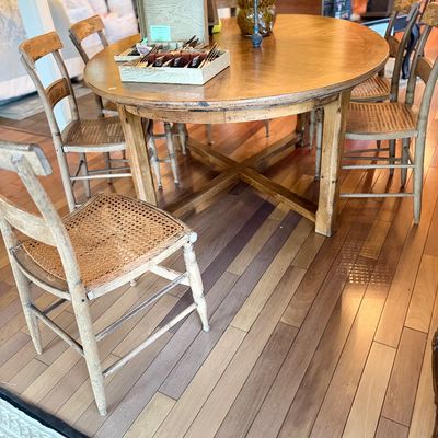 Rustic round wooden dining table with cross-base support, paired with matching cane-seat dining chairs showing distressed whitewashed...