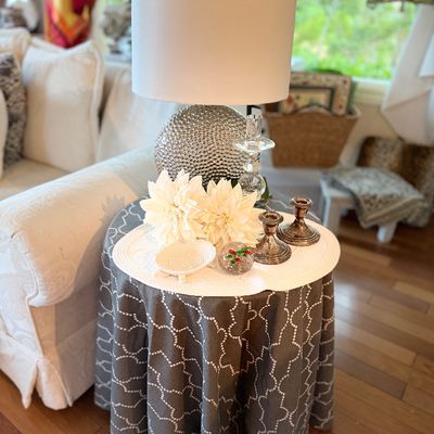 Round side table draped in grey and white patterned fabric, topped with a modern textured silver table lamp, decorative candlesticks, and...