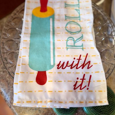 Folded kitchen towel featuring a rolling pin graphic with the phrase "Roll with it!" displayed over a clear glass plate and...
