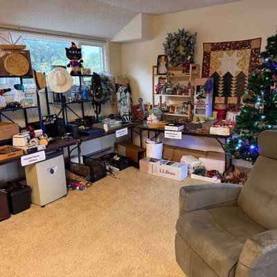 Living area display featuring seasonal holiday decorations, a lit Christmas tree, folding tables laden with household items, and a grey...