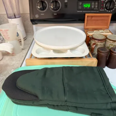 Sale Photo Thumbnail #69: Collection of kitchen housewares featuring a green heat-resistant oven mitt, plastic measuring cups, a white cake turntable, and vintage stoneware mugs.