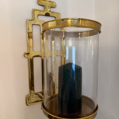 Sale Photo Thumbnail #41: Wall-mounted brass hurricane sconce featuring a geometric backplate and a clear glass cylinder shade holding a dark pillar candle.